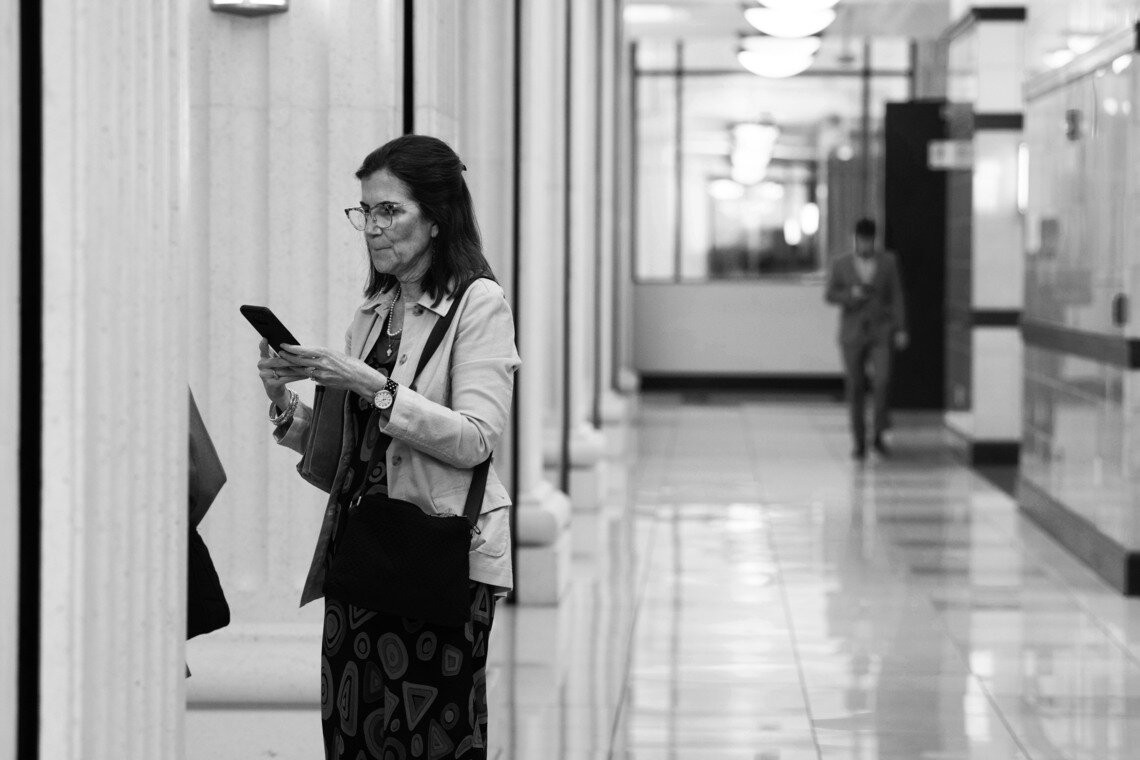 A White woman, holding her phone with both hands, stands in the hallway of a courthouse. 