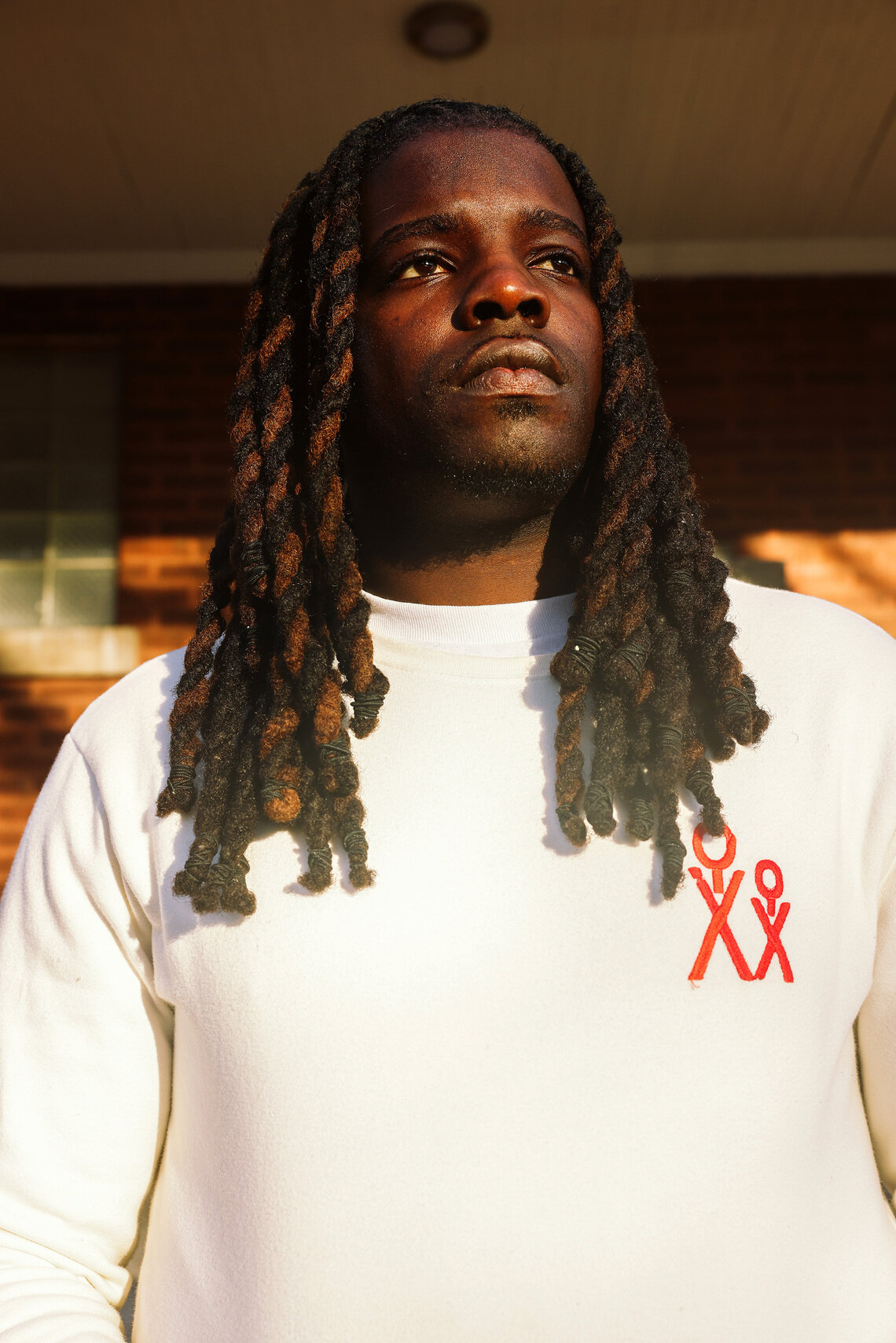 A Black man, wearing a white long-sleeved shirt, is outside on a porch and looking up to the sky. 