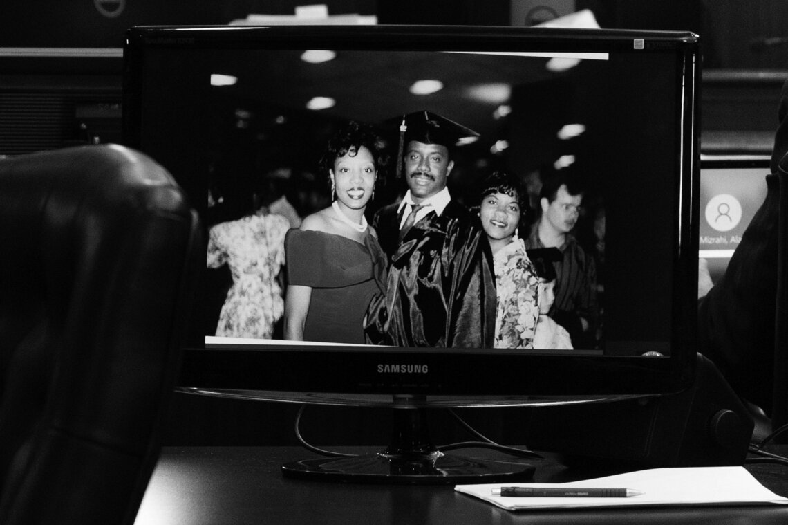 A black and white image of two Black women in dresses and a Black man in a graduation cap and gown is displayed on a courtroom monitor.