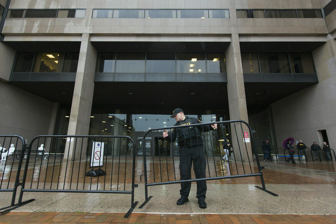 A man dressed in a black uniform and baseball cap lifts a barricade gate in front of a tall building made of reddish-brown stone.  