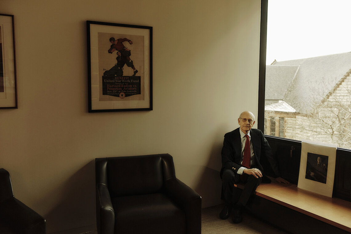 An older White man wearing glasses, a black suit, striped shirt and red tie, sits on a bench in the corner of his office. 