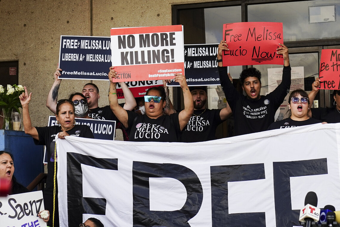 Outside of a building, a crowd representing mixed genders, ethnicities and ages stands holding signs above their heads with their mouths open. Some of the signs read “No more killing!” and “Free Melissa Lucio.” 