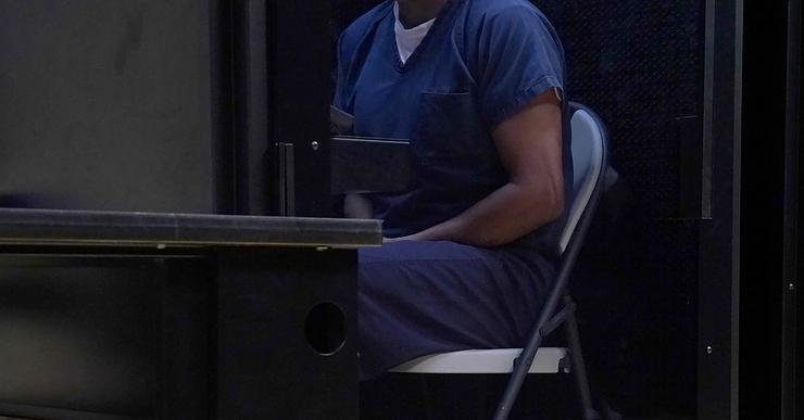 A man wearing a blue uniform sits on a metal folding chair in a small room. 